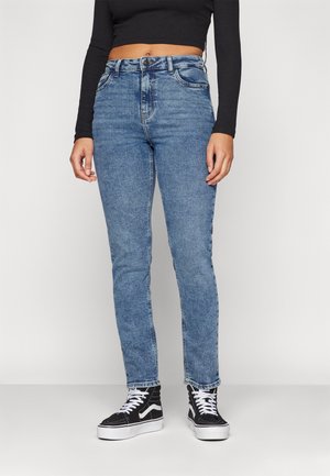 Person trägt hellblaue Skinny-Jeans mit hoher Taille, schwarzen langärmeligen Crop-Top und schwarze High-Top-Sneaker mit weißen Schnürsenkeln und Sohlen.