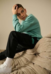 Turkis sweatshirt og sorte joggers med grønne accenter, siddende på en struktureret beige overflade, hvide sneakers, ser eftertænksom ud.