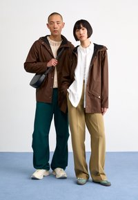 Vestes imperméables brunes avec des accents noirs, pull en tricot crème, pantalon ample vert et beige, et chaussures décontractées sur un fond bleu clair.