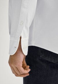 Camicia bianca con tessuto a trama, caratterizzata da un polsino arrotolato e dettaglio con bottoni. Abbinata a jeans scuri, offre un aspetto casual.