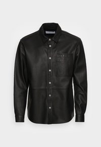 Giacca camicia in pelle nera con colletto classico, chiusura a bottoni automatici e tasca frontale con dettaglio "YPS" in rilievo. Texture liscia.