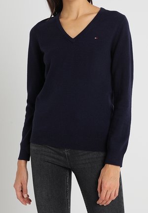 Kobieta ubrana w dopasowany granatowy sweter z dekoltem w serek i długimi rękawami oraz ciemnoszare dżinsy na jednolitym tle.