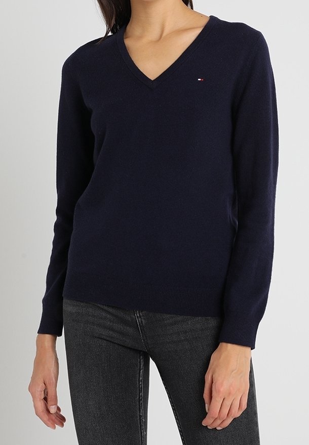 Femme portant un pull ajusté bleu marine à col en V avec manches longues et un jean gris foncé, sur un fond uni.