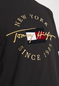 Černé bavlněné tričko s zlatou výšivkou s nápisy "NEW YORK," "Tommy Hilfiger," a "SINCE 1985," spolu s barevným blokovým logem.
