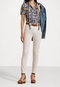 Blusa floreale con maniche corte, jeans aderenti beige chiaro, cintura marrone e sandali con zeppa marroni. Una giacca di denim è appoggiata sul braccio.
