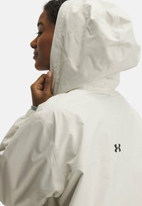 White waterproof jacket with a hood, featuring a logo on the back. The fabric has a smooth, slightly reflective texture and elastic cuffs.