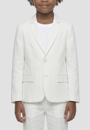 Dijete odjeveno u bijelu sako jaknu i hlače preko bijele košulje, stoji nasuprot jednobojnoj svijetlosivoj pozadini.