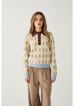 Jauna moteris dėvi gelsvos ir rudos spalvos argilo rašto megztinį su mėlynomis rankogaliais ir apykakle, dėvi laisvus šviesiai rudus kelnes, stovi priešais vienodas fonas.