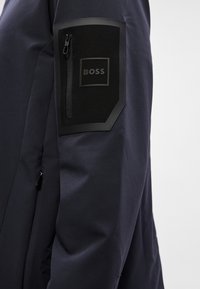 Temno modra jakna z gladko teksturo, ki ima črn žep z zadrgo in pravokotno obliž, vrezan z napisom "BOSS" na zgornjem delu rokava.