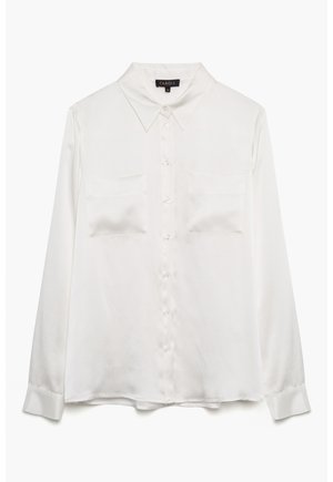 Blouse blanche à manches longues en tissu lisse avec un col pointu, des boutons sur le devant et deux poches poitrine. Les poignets sont boutonnés.