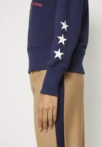 Námořnická mikina s bílými hvězdami na rukávu, kombinovaná s béžovými kalhotami s tmavě modrým postranním pruhem. Hladká struktura látky.