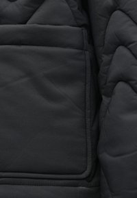 Pikowana czarna tkanina kurtki o gładkiej fakturze, z przeszywaną kieszenią i abstrakcyjnymi wzorami w pikowaniu.