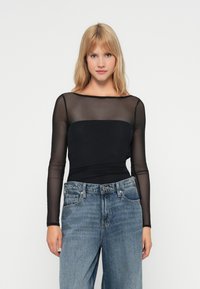 Μαύρο μπλουζάκι μακρυμάνικο με ύφασμα διχτυού και σικ λαιμόκοψη. Η εφαρμογή είναι στενή, συνδυασμένη με τζιν ψηλόμεσα σε ανοιχτό μπλε.