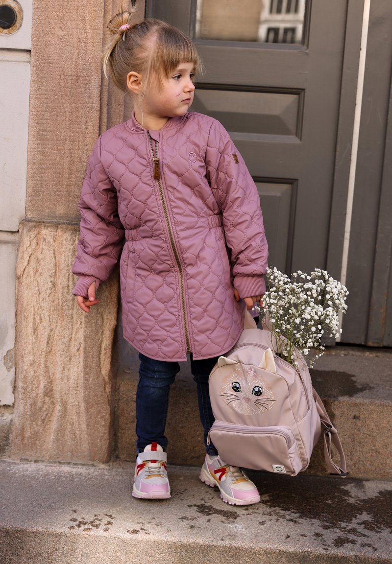 Veste matelassée rose avec une fermeture éclair, jean bleu et baskets blanches. Sac à dos beige avec un motif de visage de chat, tenant des fleurs blanches.