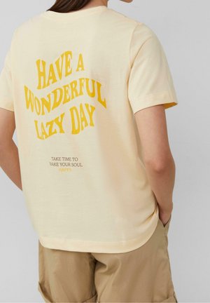 Osoba nosząca jasnożółtą koszulkę z żółtym tekstem "Have a wonderful lazy day" i mniejszym czarnym tekstem poniżej na plecach, w połączeniu z beżowymi szortami.