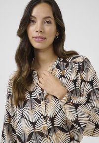 Schwarze Bluse mit einem Schalenmuster in Weiß, Beige und dezentem Braun, mit Knopfverschluss und gefältelten Details an den Schultern.