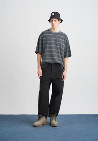 Camiseta a rayas grises de mangas cortas, pantalones vaqueros negros de corte holgado y botas beige; combinada con un gorro de pescador negro. Fondo plano.