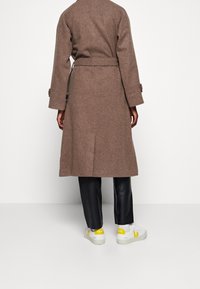 Brun uldfrakke med bælte, tætsiddende pasform, knapmanchetter; parret med sorte læderbukser og hvide sneakers med gule detaljer.