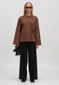 Braunes langärmliges Oberteil mit Krawatten-Detail an der Seite, kombiniert mit schwarzen weiten Hosen. Model hält eine schwarze Clutch und trägt Sonnenbrillen.
