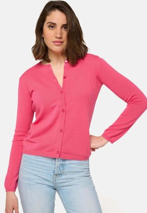 Cardigan tricoté rose avec un devant à boutons, des manches longues, des poignets côtelés et une texture douce. Associé à un jean bleu clair.