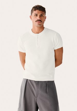 Moški s kratkimi lasmi in brki, oblečen v belo rebrasto henley majico s kratkimi rokavi in sive plisirane hlače, stoji pred enobarvnim ozadjem.