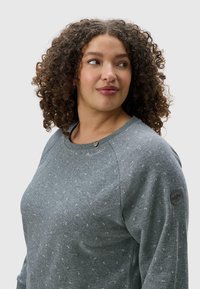 Felpa grigia con tessuto texturizzato e piccole macchie multicolori. Presenta un collo rotondo, maniche raglan e un'emblema decorativa sulla spalla.