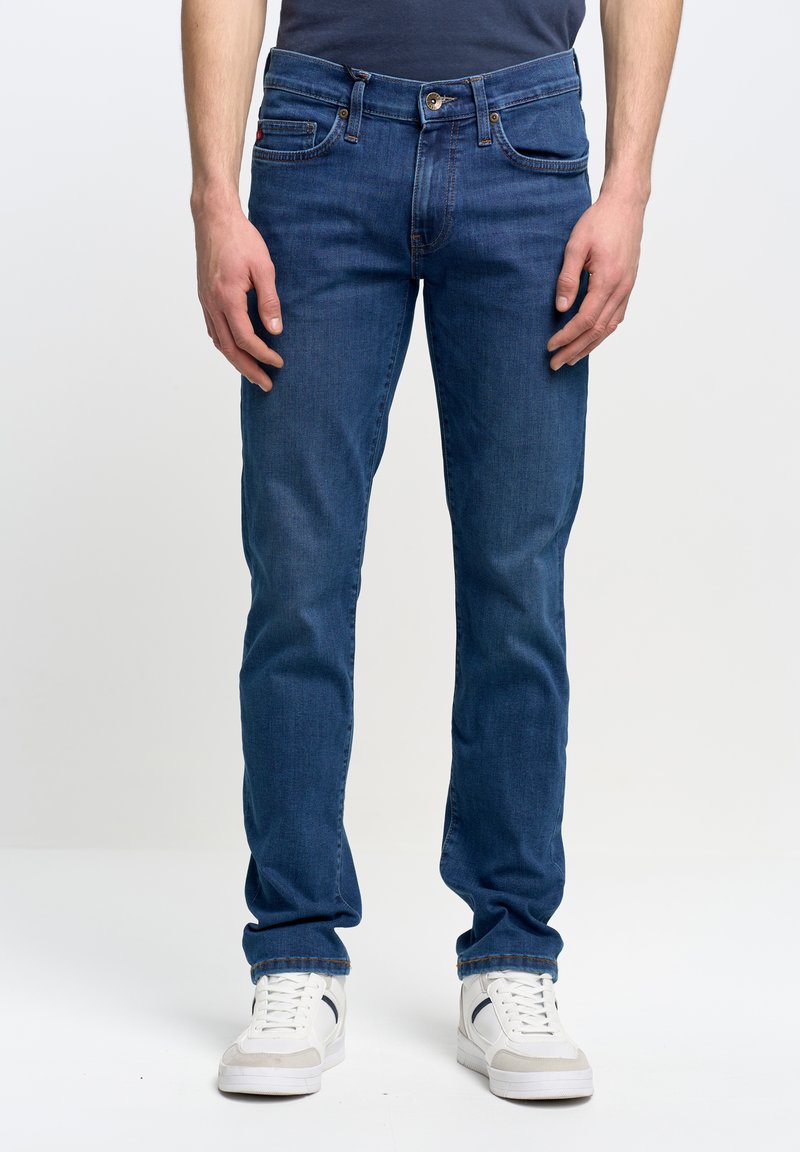 Mees, kes kannab siniseid sirge lõikega teksapükse ja valgeid tenniseid, seisab ühevärvilise valge tausta ees.