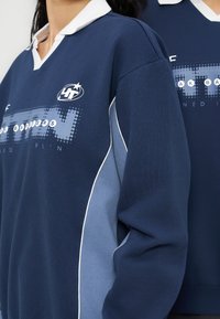 Δύο άτομα που φορούν μπλε σκούρα και ανοιχτά μπλε μακρυμάνικα πουκάμισα με γιακά και σχέδιο "4T" και κείμενο στο στήθος.