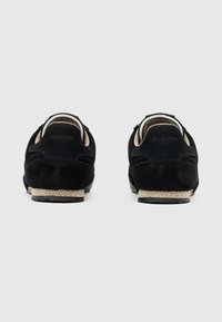 Schwarze Wildleder-Sneaker mit texturierter Oberfläche, kontrastierender beiger Gummisohle und geprägtem Branding an der Ferse. Schlichtes, elegantes Design.