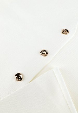 Witte textuurrijke stof met drie onregelmatige ronde gouden knopen diagonaal gerangschikt op een opgevouwen kledingstuk.