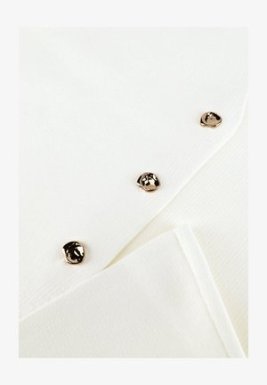 Witte textuurrijke stof met drie onregelmatige ronde gouden knopen diagonaal gerangschikt op een opgevouwen kledingstuk.