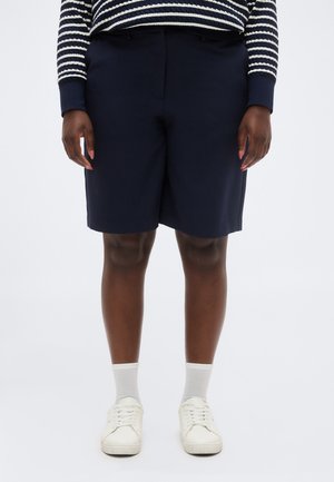 Personne portant un short bleu marine jusqu'aux genoux, un shirt à manches longues rayé, des chaussettes blanches et des baskets blanches, debout devant un fond uni.