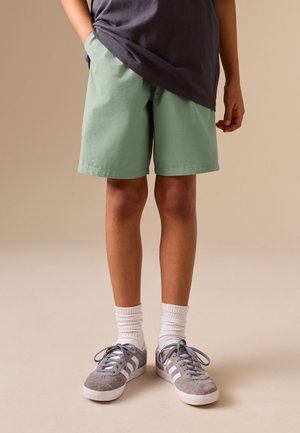 Kind trägt hellgrüne Shorts, graue Sneakers mit weißen Streifen, weiße Socken und ein dunkelgraues Shirt mit einer Hand in der Tasche.