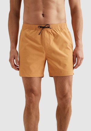 Mužský trup a nohy v horčicovožltých plavkách s elastickým pásom a čiernym šnúrkou, stojaci na neutrálnom pozadí.