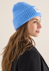 Niebieska prążkowana dzianinowa czapka z podwiniętym mankietem i prostokątną białą metką z czarnym napisem. Miękka w dotyku, dopasowany kształt.