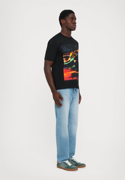 Man standing wearing a black graphic tee with abstract bright colors, light blue jeans, and green sneakers against a plain white background.