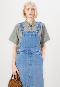 Lys blå denimkjole med for lomme og metallfester, kombinert med en grå kortermet skjorte med knapper. Brun håndveske holdt i hånden.