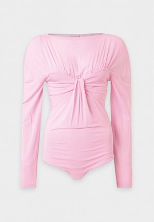 Mulher vestindo um top rosa de manga comprida com nó à frente e jeans azuis de perna larga com cinto amarrado, em pé com as mãos nos bolsos.
