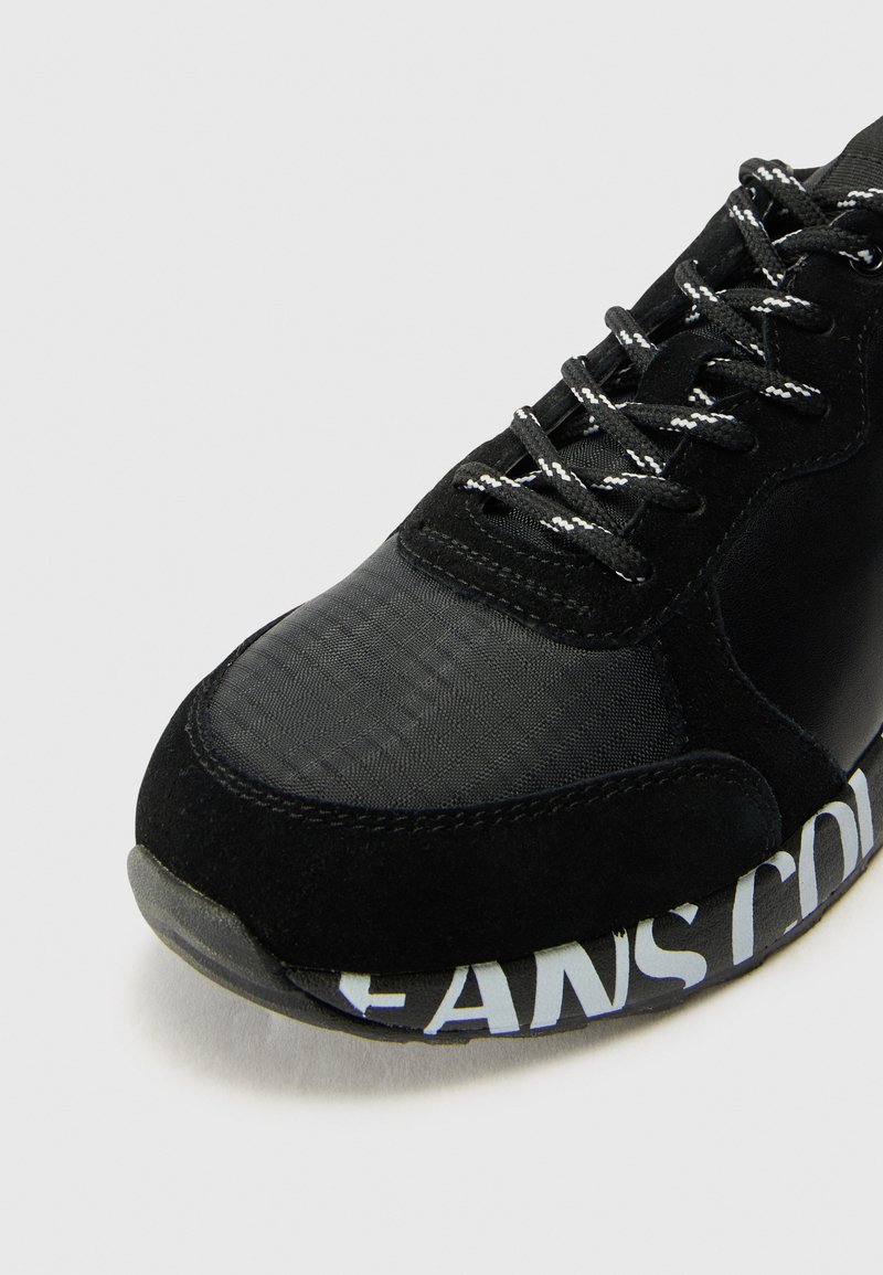 Zwarte sneaker met een mix van suede en textiel, met een ronde neus en witte tekstdetails op de tussenzool. Zwarte veters en accenten.