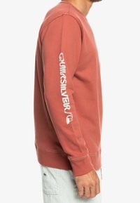 Rostfärgad sweatshirt med rund halsringning, med vit "Quiksilver"-text längs vänster ärm samt ribbade muddar och nederkant.