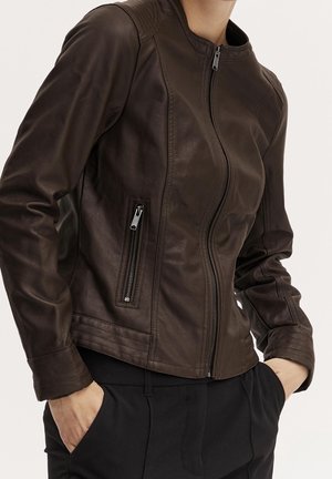 Giacca aderente in pelle marrone scuro con cerniera frontale e tasche laterali con zip indossata sopra pantaloni neri.