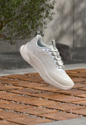 Λευκό αθλητικό παπούτσι Calvin Klein με χοντρή σόλα, κορδόνια και λογότυπο στο πλάι, αιωρούμενο πάνω από δάπεδο με τερρακότα πλακάκια και βότσαλα σε εξωτερικό χώρο.