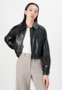 Svart fuskeskinn cropped jakke med spredt krage, glidelås og elastisk nederkant. Båret over en hvit topp og beige bukser.