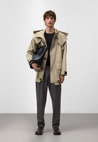 Beige arbetsjacka med stor huva, knappad framtill och fickor. Svart väska hålls under armen. Grå byxor och mörka skor avslutar looken.