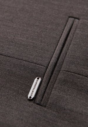 Gros plan sur un tissu chevrons gris foncé avec une poche passepoil verticale et une petite étiquette métallique marquée « QUINTESSENCE ».
