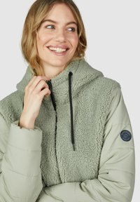 Veste verte avec un devant en polaire duveteuse et des manches en nylon lisse ; elle comprend une capuche, une fermeture éclair et un logo rond sur la manche.