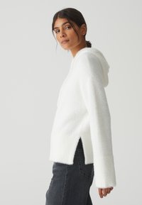 Weißer, flauschiger Hoodie mit lässiger Passform, seitlichen Schlitzen, langen Ärmeln und weichem Material, getragen über dunkelgrauen Jeans.