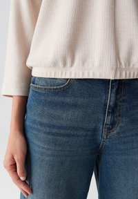 Hellbeige, geripptes Oberteil mit gerafftem Saum, kombiniert mit blauen Jeans aus Denim mit geschwungenen Vordertaschen und kontrastierenden Nähten.