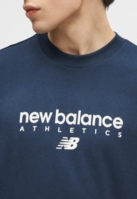 Άνδρας  που φοράει μπλε σκούρο φούτερ με λευκή επιγραφή «new balance ATHLETICS» και λογότυπο NB στο μπροστινό μέρος, κοιτάζοντας πλευρικά.