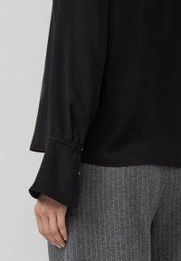 Zwarte blouse met een relaxte pasvorm, voorzien van uitlopende manchetten en een gladde textuur, gecombineerd met grijze broek met streepjes.